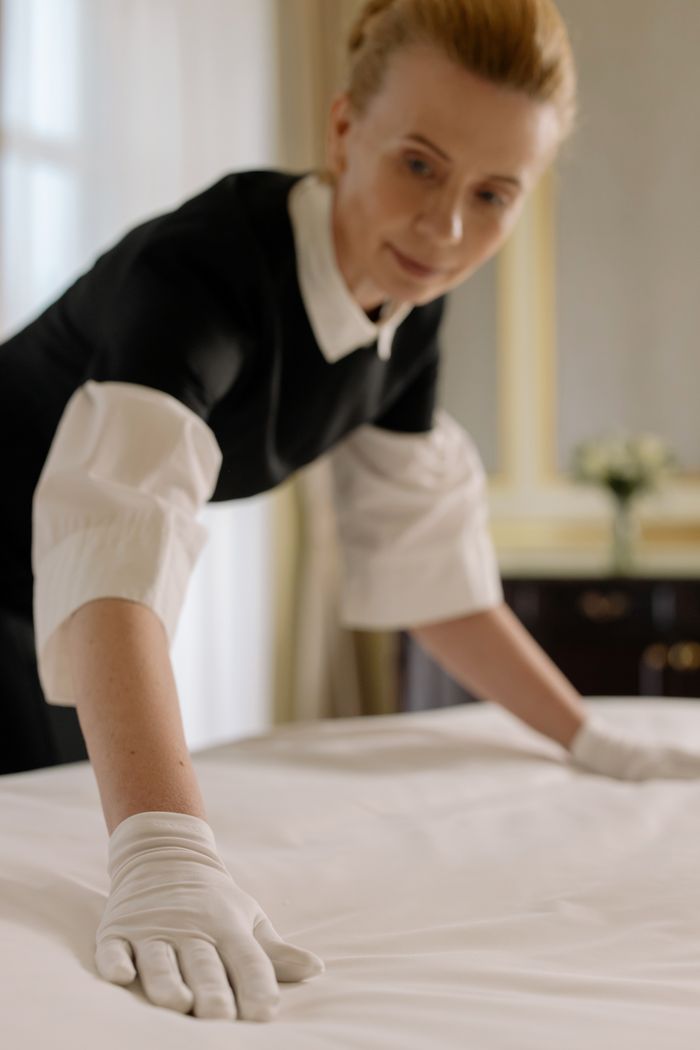 In a refined hotel room, a housekeeper gracefully straightens the bed linens, highlighting skilled hospitality service.